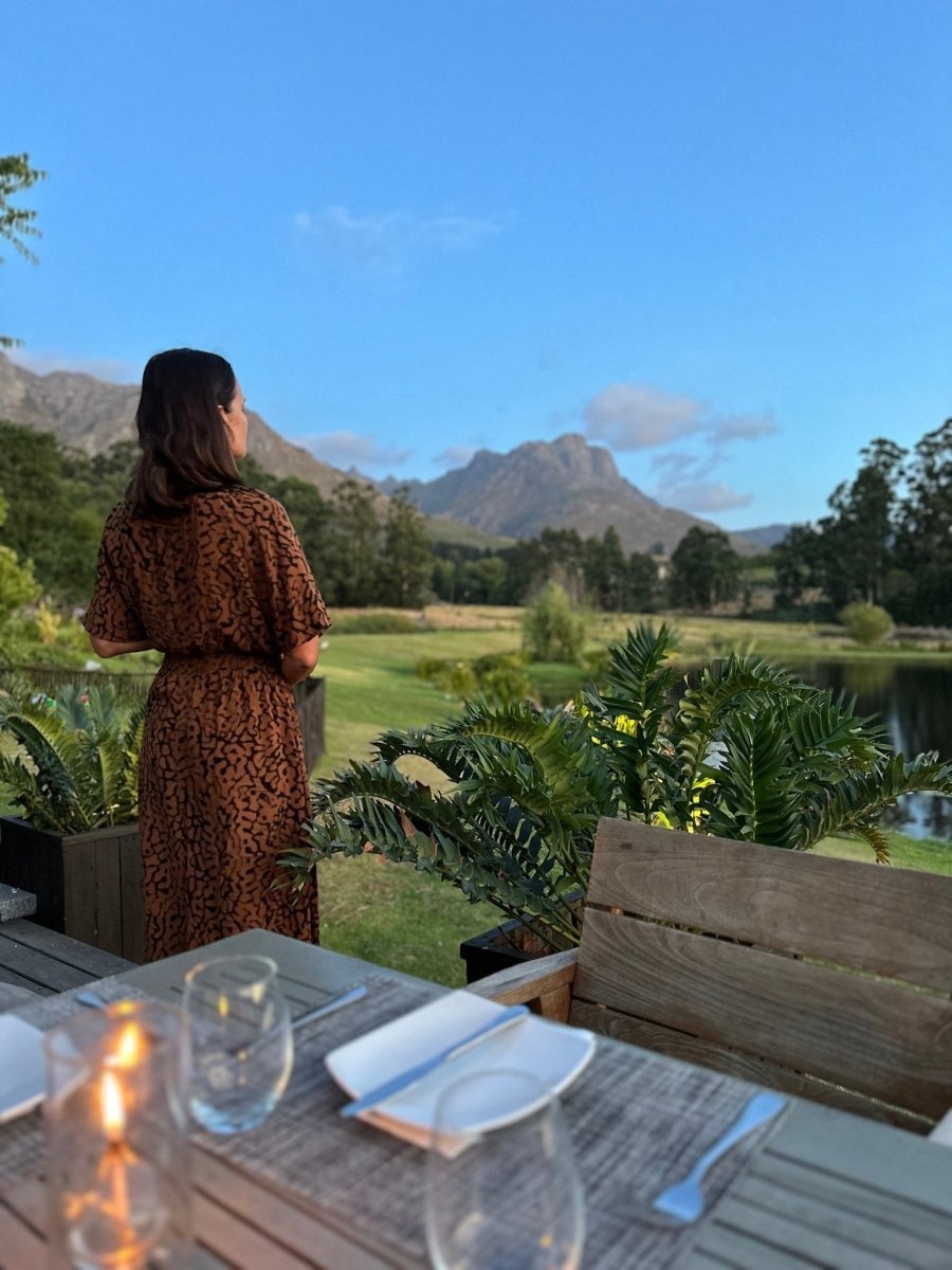 Eine Frau in einem braun gemusterten Kleid steht auf einer Holzterrasse und blickt auf Berge und einen Teich. Der kerzenbeleuchtete Esstisch ist perfekt für Reisefotografie-verbessern Sie Ihre Fotos mit dem kidswelltraveled mobile Preset Lightroom von kidswelltraveled.