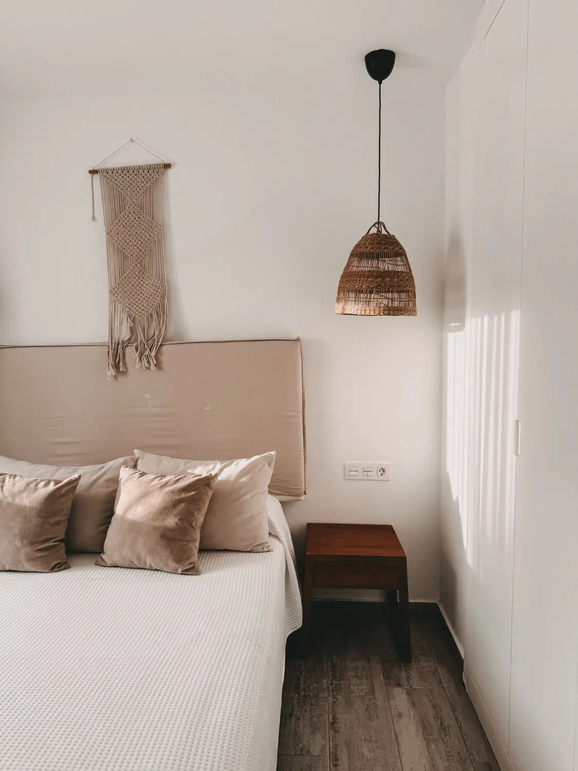 Minimalistisches Schlafzimmer im Shambhala Fuerteventura - Ein Wohlfühlort für Eltern und Kinder auf den Kanaren von kidswelltraveled, mit beigen Kissen, einem Nachttisch aus Holz, einer geflochtenen Pendelleuchte, Makramee-Wanddekoration und sonnenbeschienenen Schranktüren.