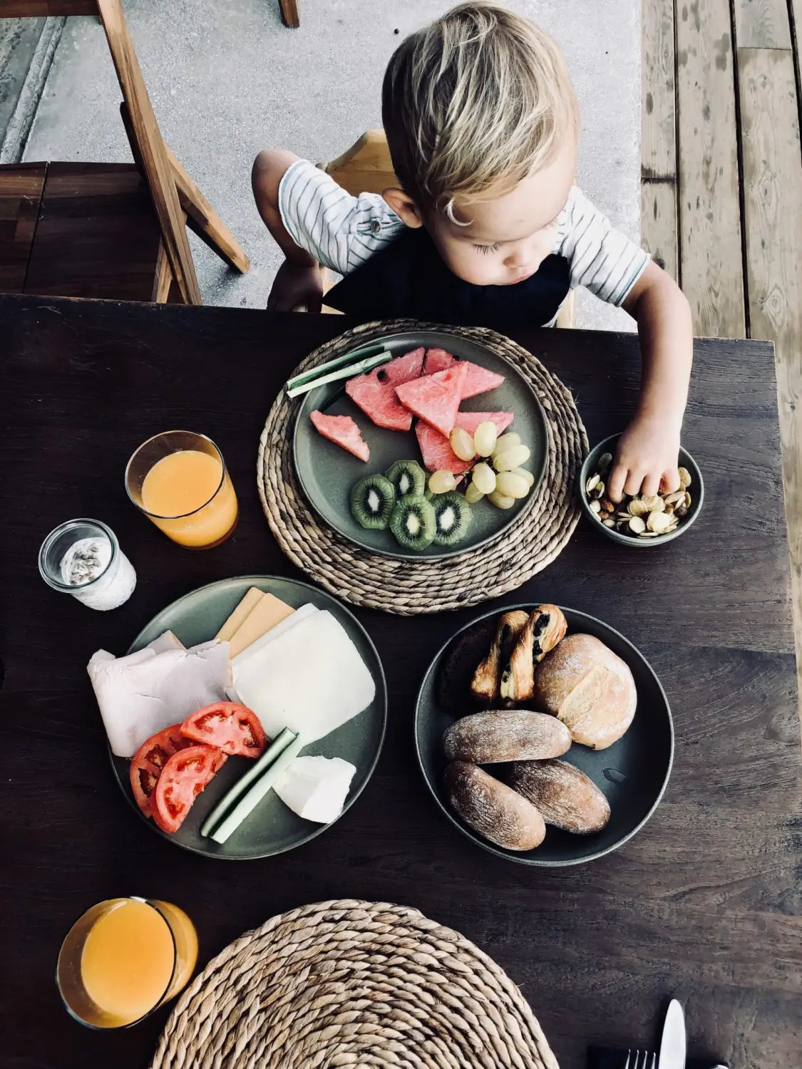 A small child enjoys fresh fruit, cheese, sausages, bread rolls and nuts at a dark wooden table in Das Domes Zeen in Crete by kidswelltraveled - an inviting boho family hotel with orange juice and woven placemats.