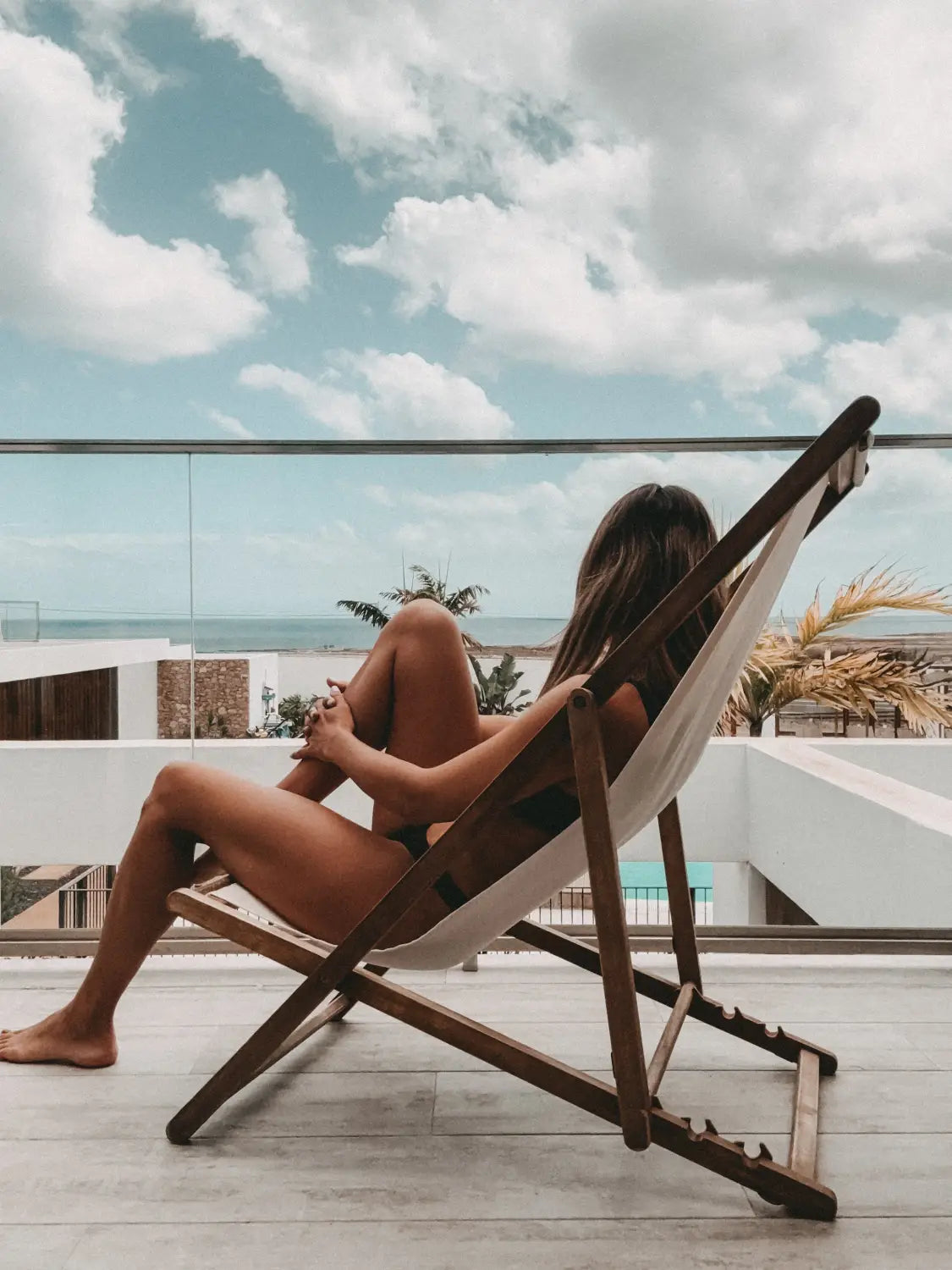 Eine Person entspannt sich auf einem Holzsessel im Shambhala Fuerteventura - Ein Wohlfühlort für Eltern und Kinder auf den Kanaren von kidswelltraveled, mit Meerblick, Glasgeländer, Palmen und moderner Kulisse - perfekt für Familienurlaub.