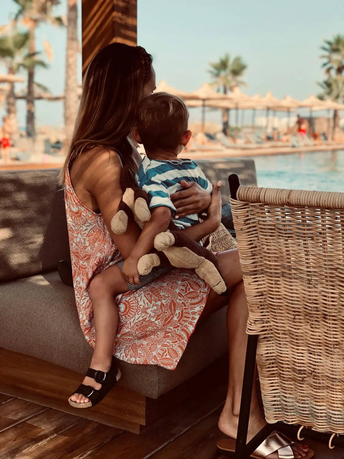 At Das Domes Zeen in Crete by kidswelltraveled, a woman in a patterned dress sits with child and toy monkey on a bench, looking towards the sunny pool area with palm trees and loungers - a boho dream for families.