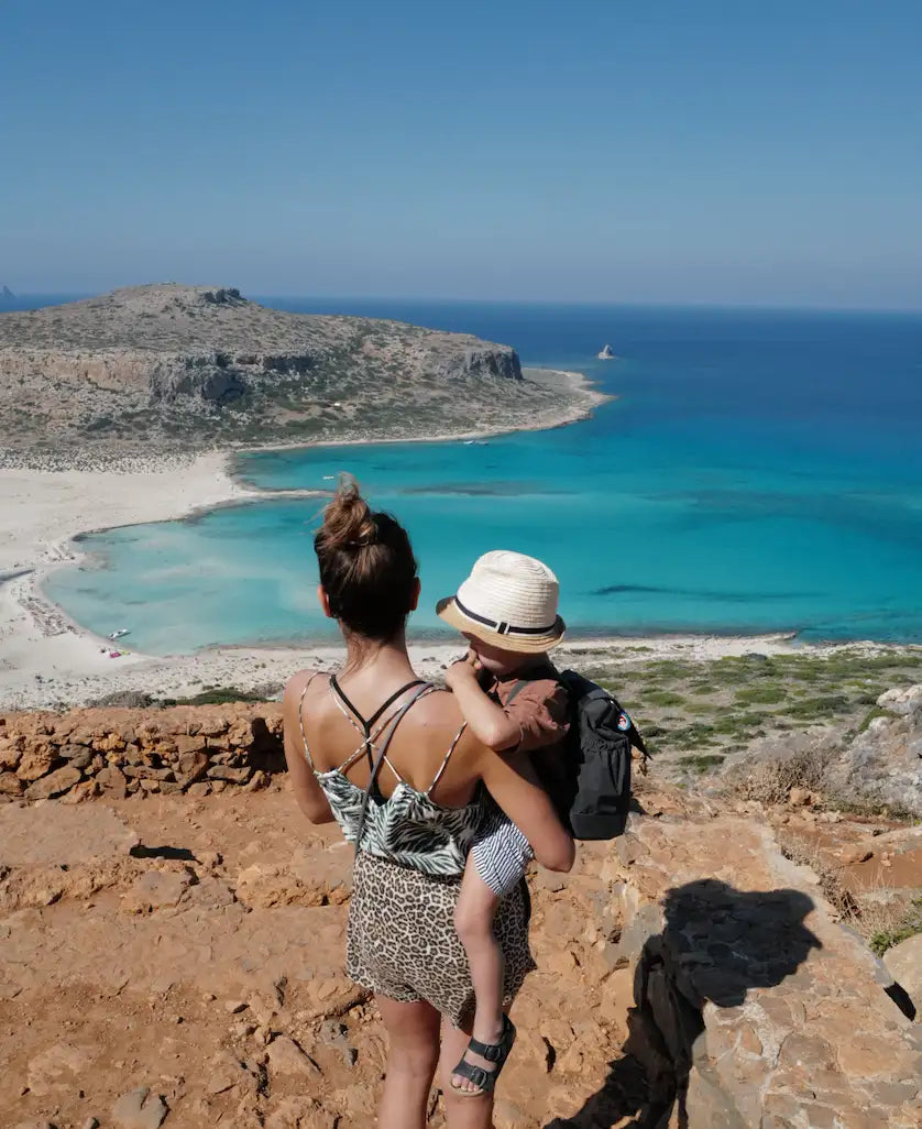 Eine Frau mit einem kleinen Kind im Arm überblickt von einem felsigen Aussichtspunkt aus eine türkisfarbene Bucht und einen Sandstrand, während im Hintergrund der blaue Himmel und die zerklüftete Küste zu sehen sind.