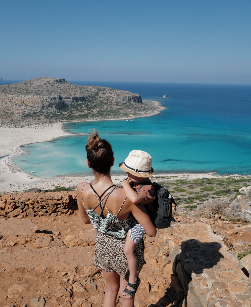 Eine Frau mit einem kleinen Kind im Arm überblickt von einem felsigen Aussichtspunkt aus eine türkisfarbene Bucht und einen Sandstrand, während im Hintergrund der blaue Himmel und die zerklüftete Küste zu sehen sind.