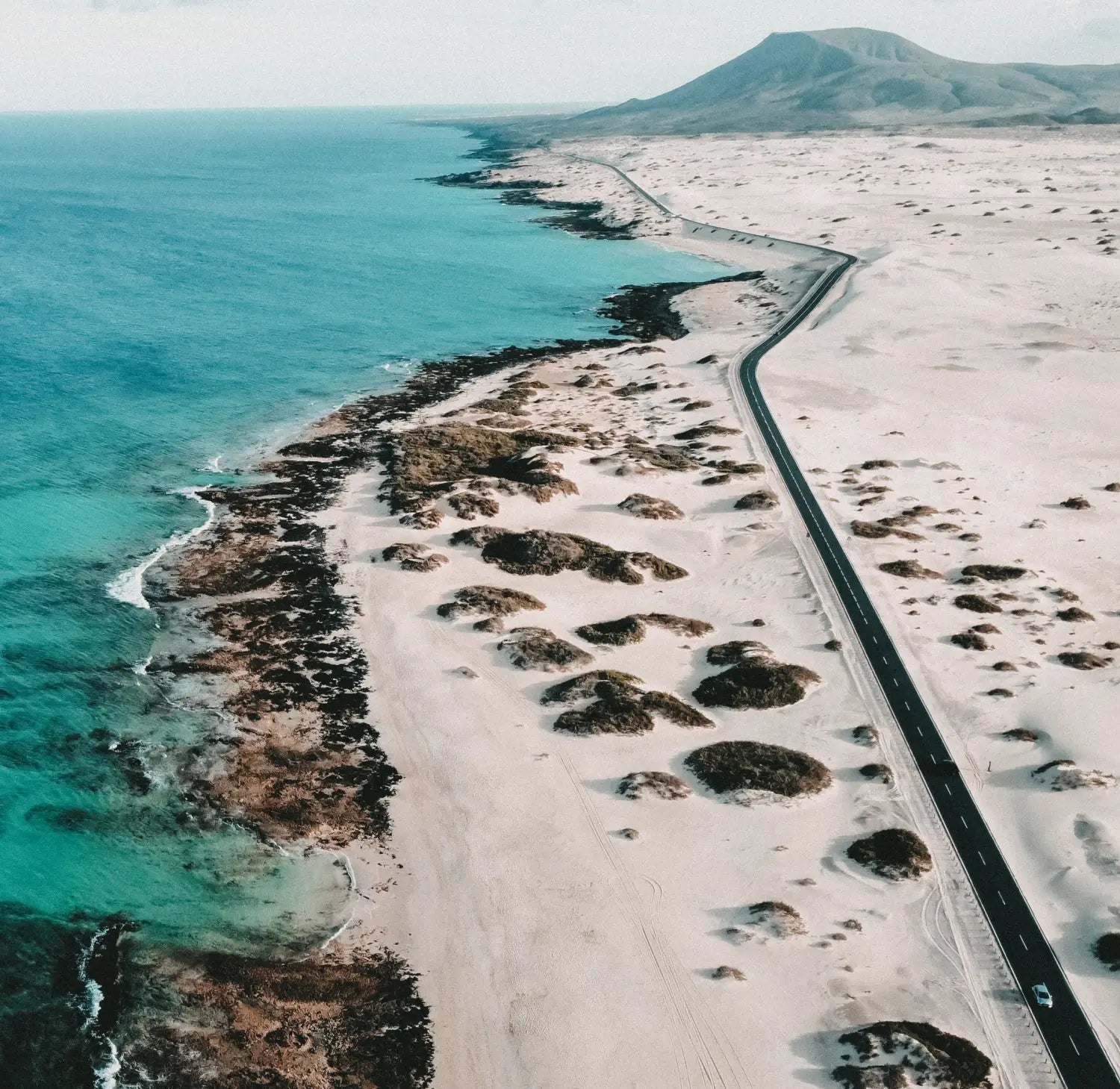 Ein Küstenluftbild in der Nähe von Shambhala Fuerteventura - Ein Wohlfühlort für Eltern und Kinder auf den Kanaren von kidswelltraveled zeigt kurvenreiche Straßen, weiße Dünen und türkisblaues Meer-ideal für Familienurlaub unter klarem Himmel.