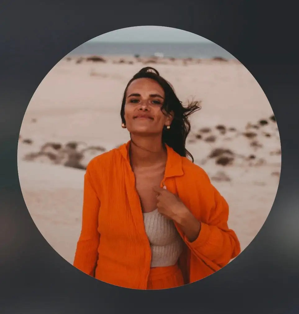 Eine Frau steht an einem Sandstrand und trägt ein orangefarbenes Outfit über einem leichten Oberteil. Sie hat langes dunkles Haar, lächelt sanft, und im Hintergrund sind Dünen und ein bewölkter Himmel zu sehen.